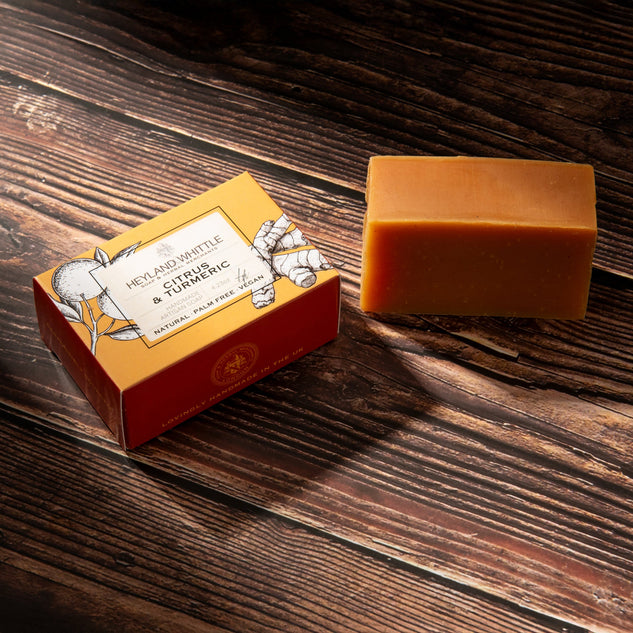 A rectangular bar of orange-amber soap sits beside its branded orange and red box on a dark wood surface. The box reads 'Heyland & Whittle Ginger & Turmeric Natural Plant Based Soap.