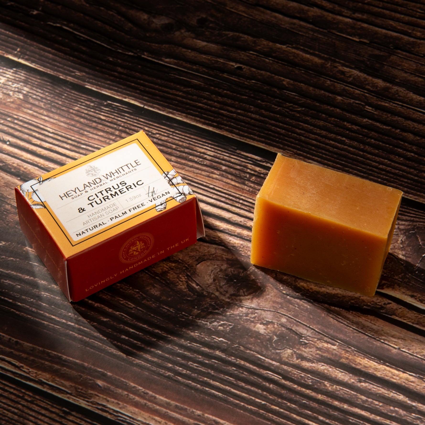 A rectangular bar of orange-yellow soap sits beside a small branded box on a dark wooden surface. The box reads Heyland Whittle Citrus & Turmeric.