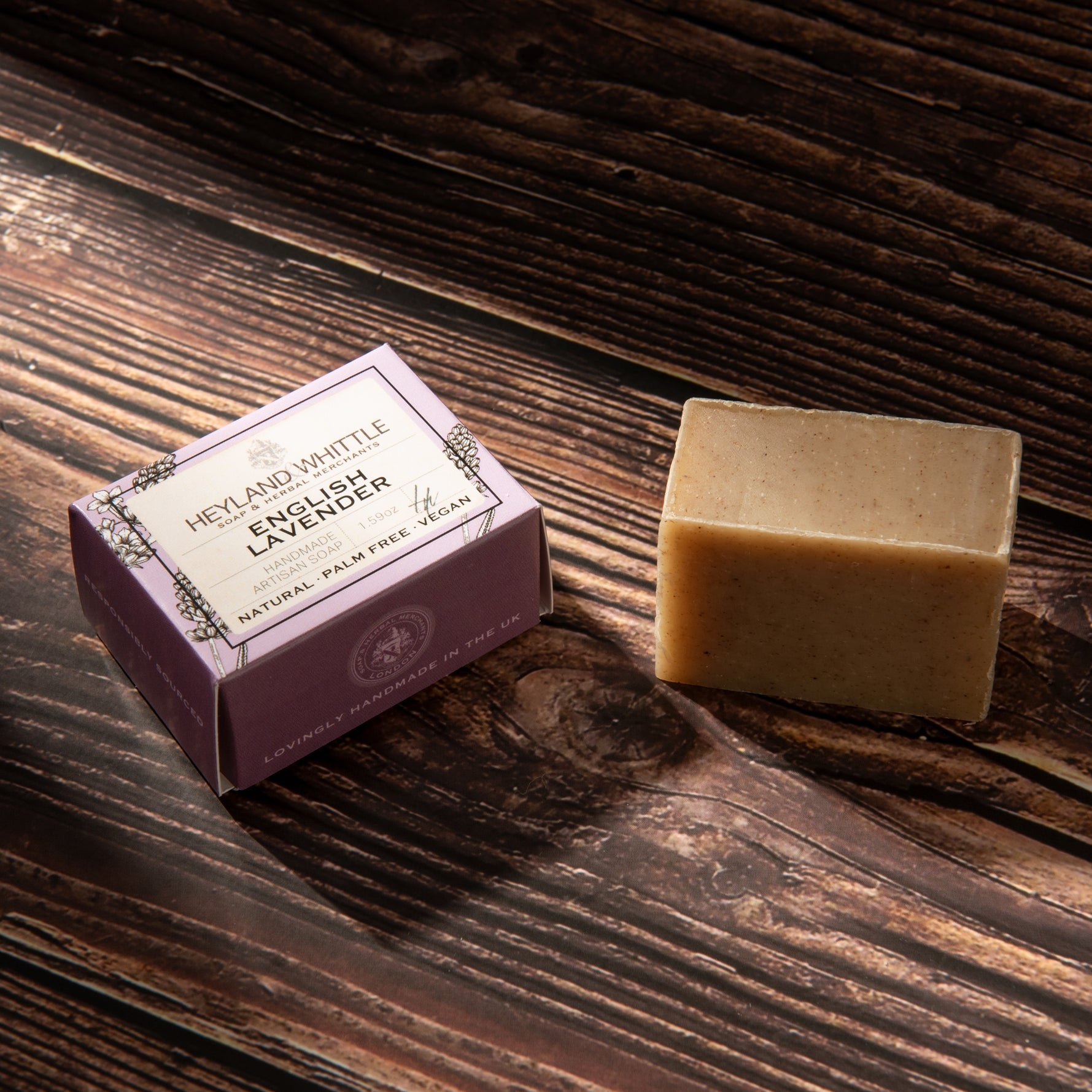 A bar of cold processed soap next to its purple packaging box on a dark wooden surface. The box is labeled 'Heyland & Whittle English Lavender'.