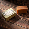 A bar of soap with dark specks sits next to its cream-colored packaging box on a dark wooden surface. 