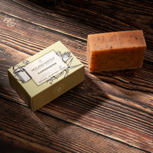 A bar of soap with dark specks sits next to its cream-colored packaging box on a dark wooden surface. 