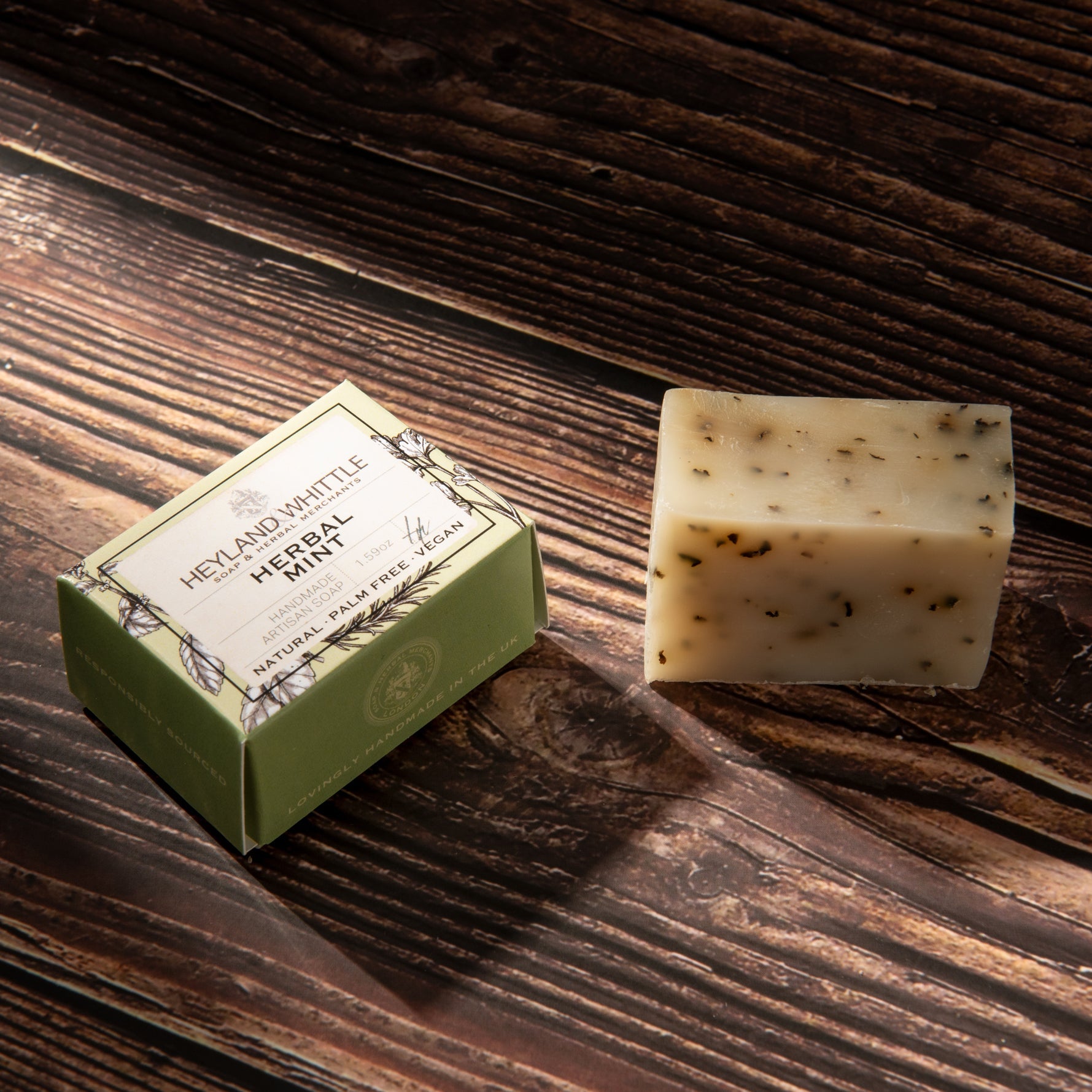 A bar of herbal mint soap next to its green paper packaging box on a dark wooden surface. 