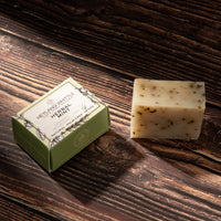 A bar of herbal mint soap next to its green paper packaging box on a dark wooden surface. 