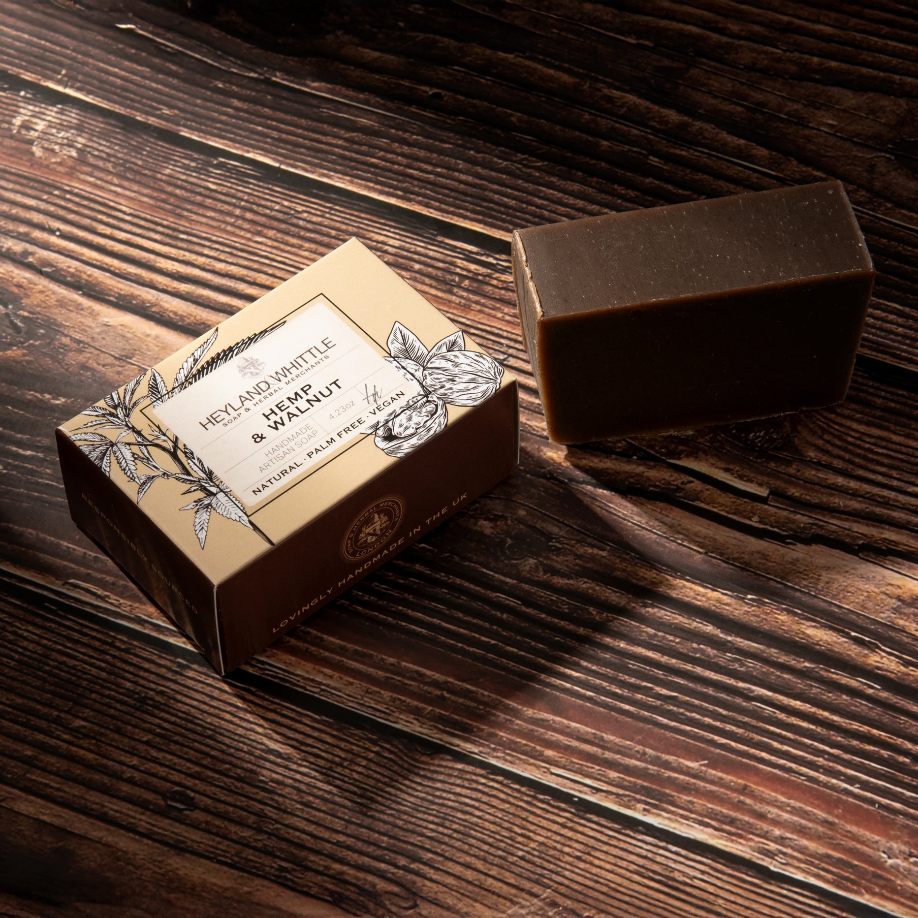 A rectangular bar of dark brown soap sits beside its decorative cardboard box on a rustic dark wood surface. 