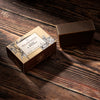 A rectangular bar of dark brown soap sits beside its decorative cardboard box on a rustic dark wood surface. 