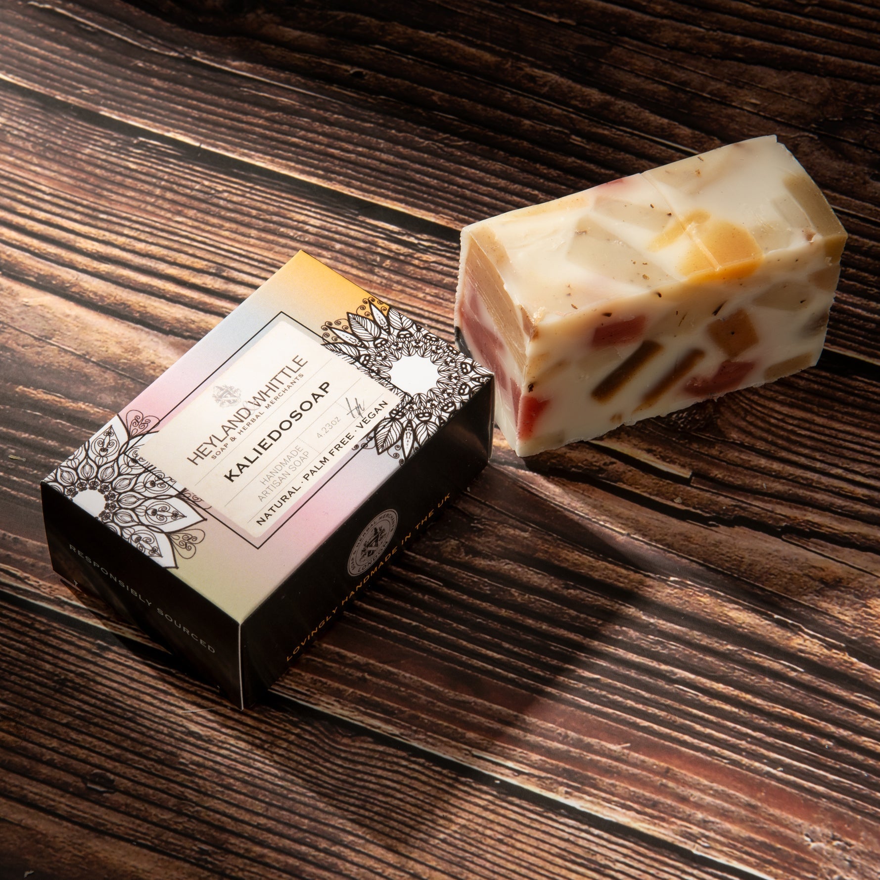 A rectangular bar of handmade soap with visible fruit pieces and flecks, placed beside its decorative retail box on a dark wood surface.
