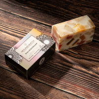 A rectangular bar of handmade soap with visible fruit pieces and flecks, placed beside its decorative retail box on a dark wood surface.