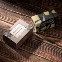A rectangular bar of marbled soap with black and cream patches sits beside its brown branded box on a dark wooden surface. 
