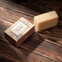 A rectangular bar of pale beige soap sits on a dark wooden surface next to its kraft paper box. 