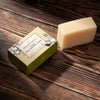 A rectangular bar of pale beige soap stands beside a small olive-green branded box on a dark wooden surface. 