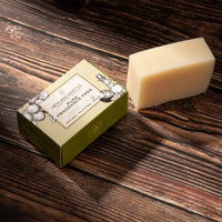A rectangular bar of pale beige soap stands beside a small olive-green branded box on a dark wooden surface. 