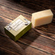 A rectangular bar of pale beige soap stands beside a small olive-green branded box on a dark wooden surface. 