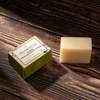 A bar of pale cream-colored soap next to its green and white packaging box on a dark wood background. The box is labeled 'Heyland and Whittle Pure Fragrance Free'.