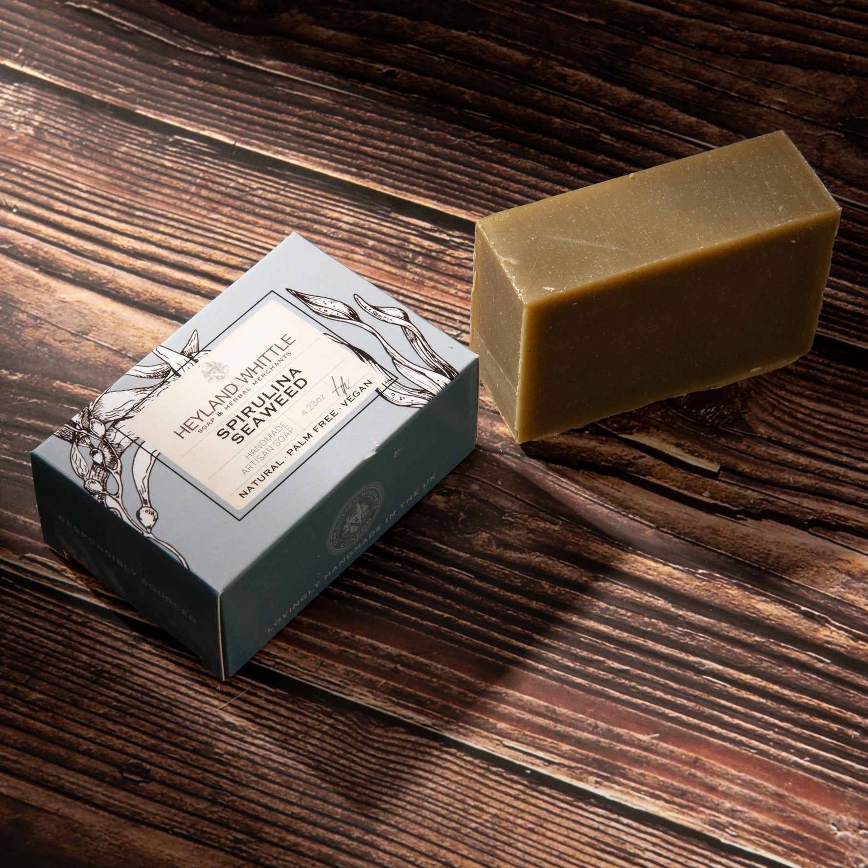 A rectangular bar of natural soap in a warm tan color sits next to its blue-gray packaging box on a dark wood background. 