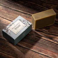 A rectangular bar of natural soap in a warm tan color sits next to its blue-gray packaging box on a dark wood background. 