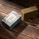 A rectangular bar of natural soap in a warm tan color sits next to its blue-gray packaging box on a dark wood background. 