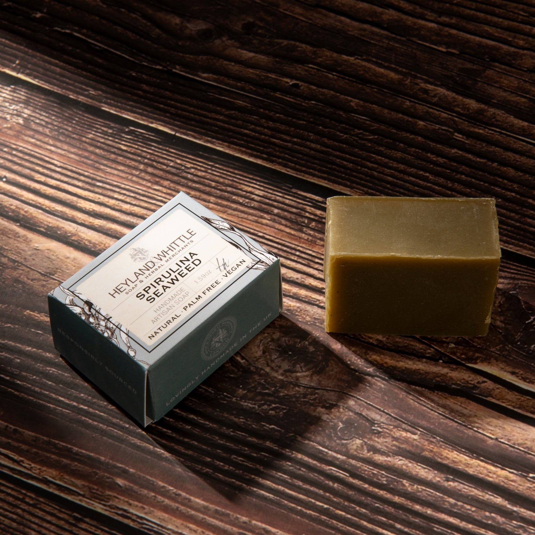 A rectangular bar of natural soap in a muted olive-green color sits next to its decorative packaging box on a dark wood background. 