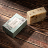 A rectangular bar of handmade soap with visible botanical inclusions sits on a dark wood surface next to its light green paper 
