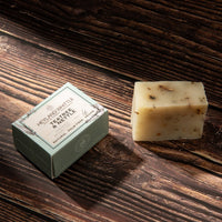 A bar of natural soap and its matching box from Heyland & Whittle, featuring the scent 'Pectree & Neille'. The soap is a pale cream-beige with visible botanical flecks, displayed next to its light sage-green packaging on a dark wood background.