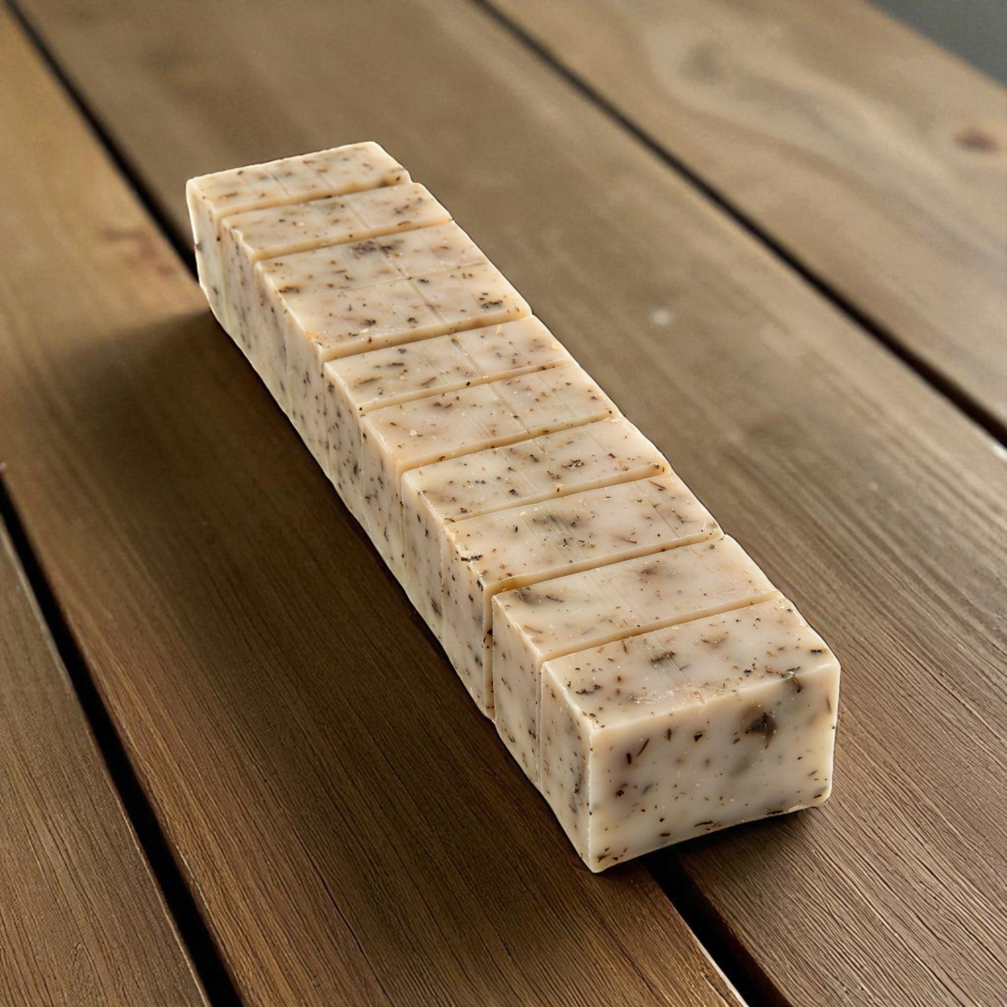 A rectangular bar of handmade soap, sliced into ten pieces, resting diagonally on a rustic wooden plank surface.