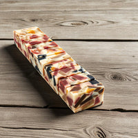 A rectangular bar of artisanal soap with a marbled pattern of cream, pink, orange, and black, sliced into small square pieces and arranged diagonally on a rustic wooden surface.