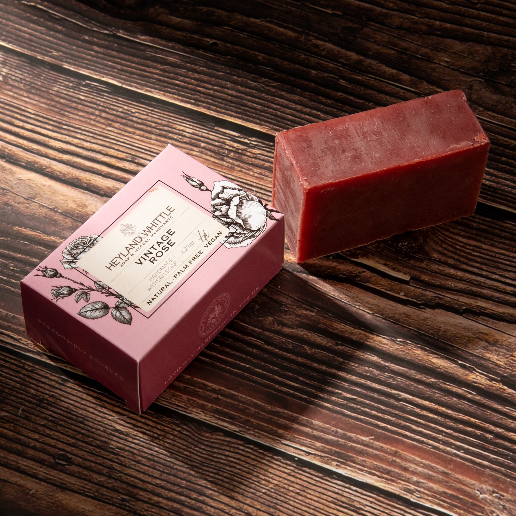 A rectangular bar of deep red soap next to its pink decorative packaging box on a dark wood background.