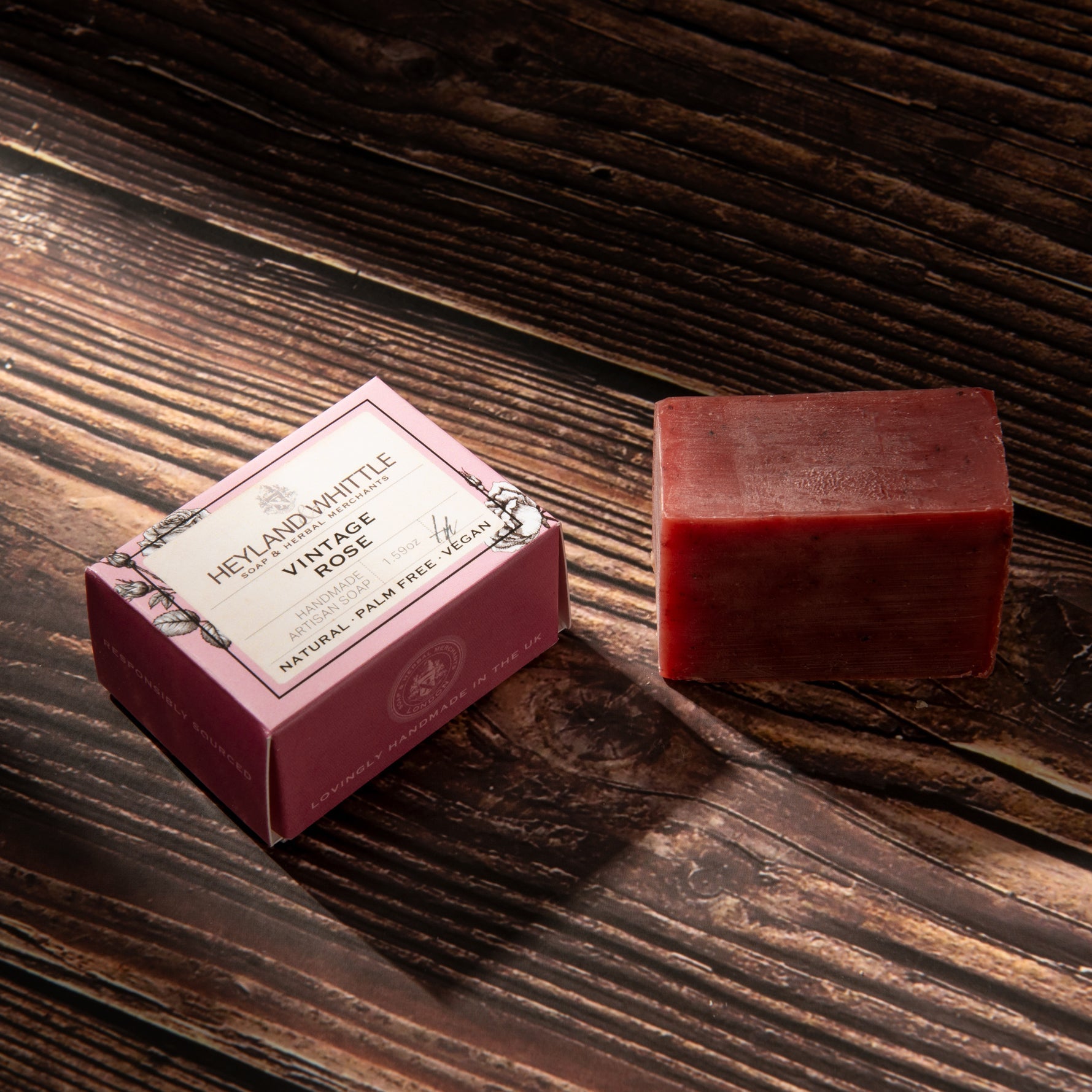 A rectangular bar of pinkish-red soap sits beside its pink box on a dark rustic wooden surface. 
