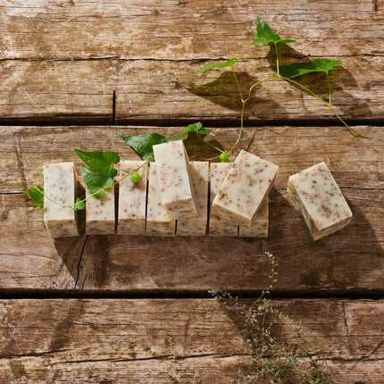 Several rectangular bars of handmade soap with visible botanical specks, arranged on a rustic wooden surface with green leaves and stems as styling props.