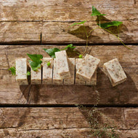 Several rectangular bars of handmade soap with visible botanical specks, arranged on a rustic wooden surface with green leaves and stems as styling props.
