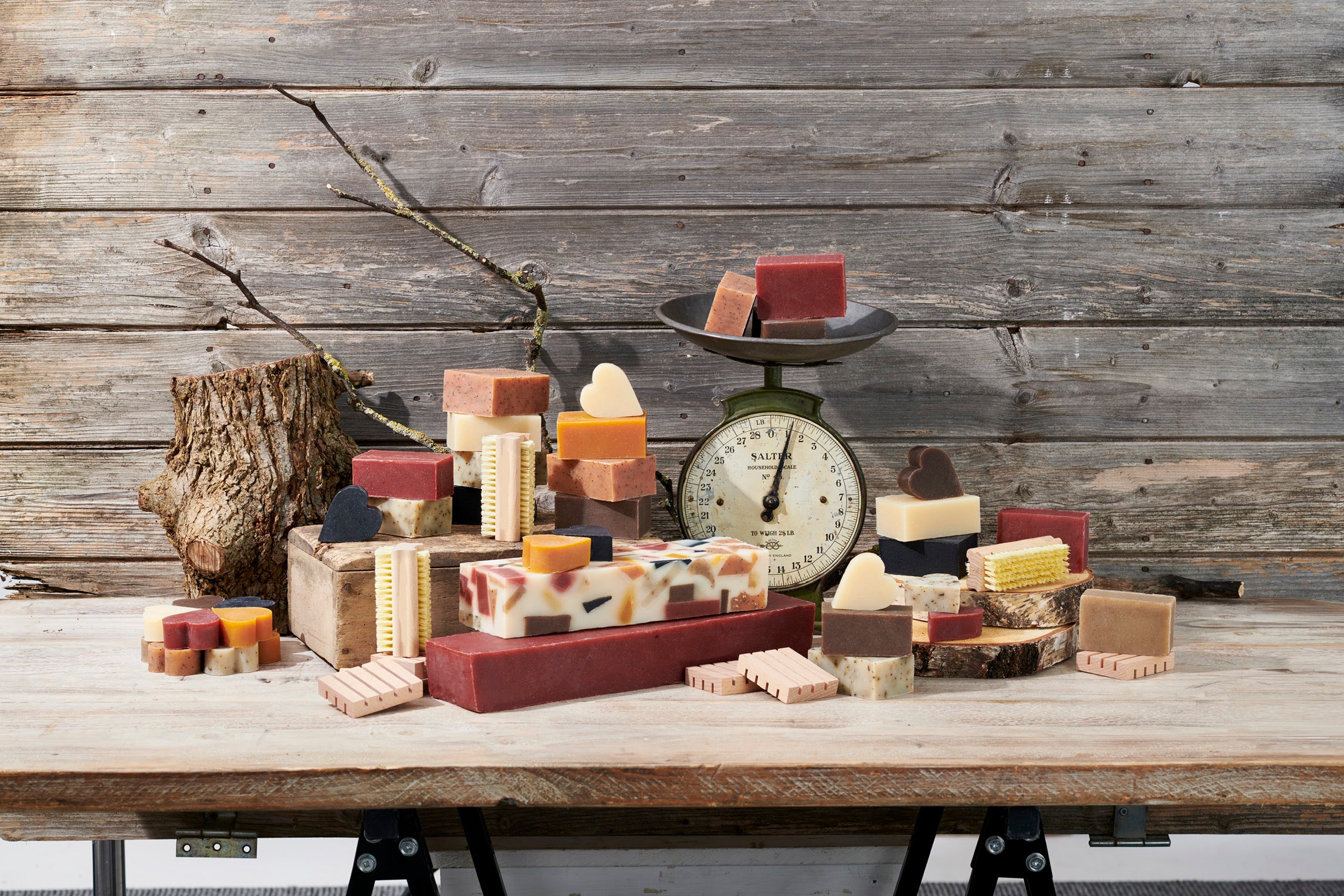 An assortment of handmade artisanal soaps and bars arranged on a rustic wooden table in front of a weathered wood plank wall. The soaps feature various colors including terracotta red, cream, yellow, and brown, with some decorated with stripes or hearts.