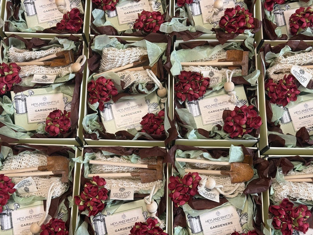 A grid arrangement of gift boxes containing gardening kits with wooden tools, lace, green leaves, and deep red hydrangea flowers. 