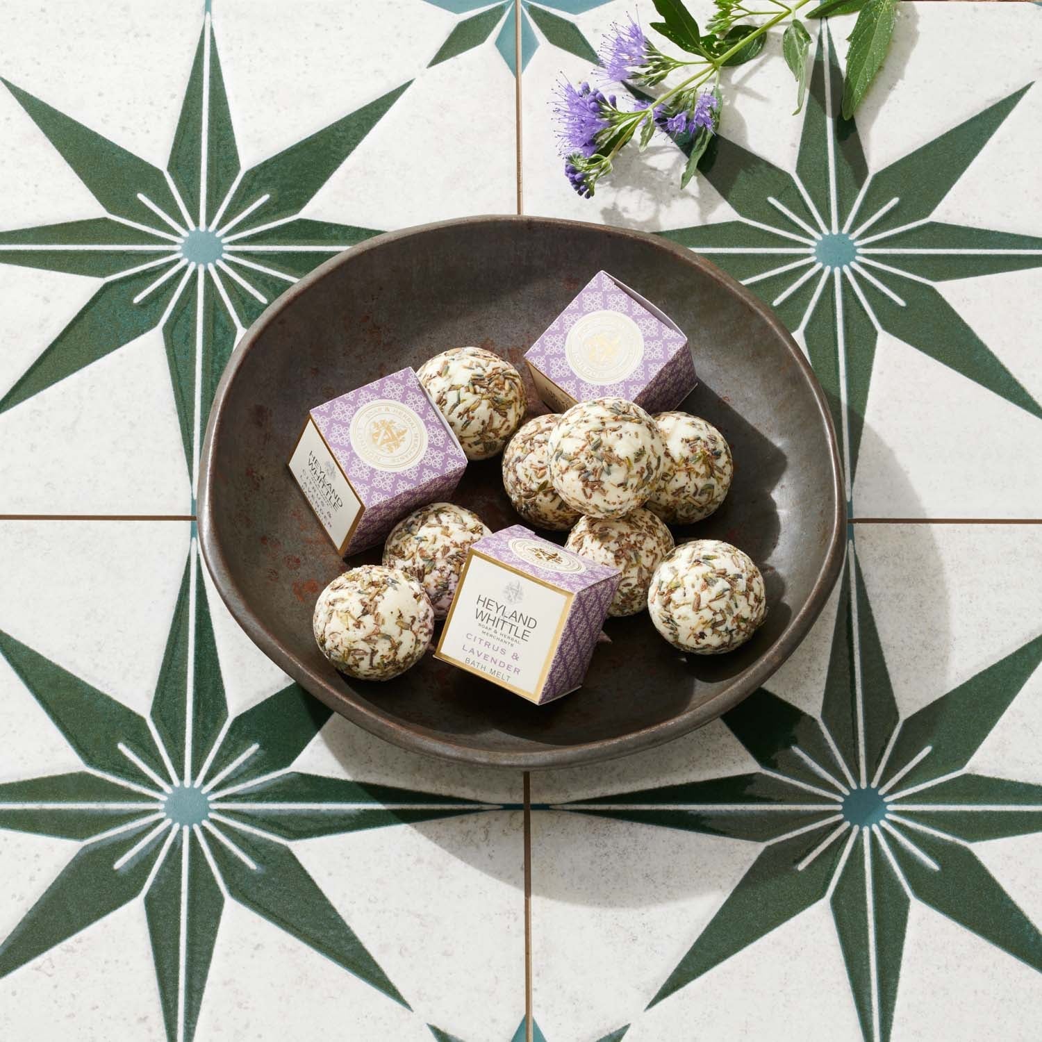 A dark bowl filled with bath melts and two small purple-and-white boxes, arranged on a decorative tile surface with starburst patterns and a sprig of purple flowers.