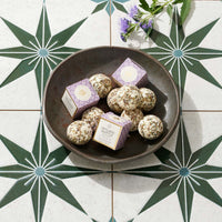 A dark bowl filled with bath melts and two small purple-and-white boxes, arranged on a decorative tile surface with starburst patterns and a sprig of purple flowers.