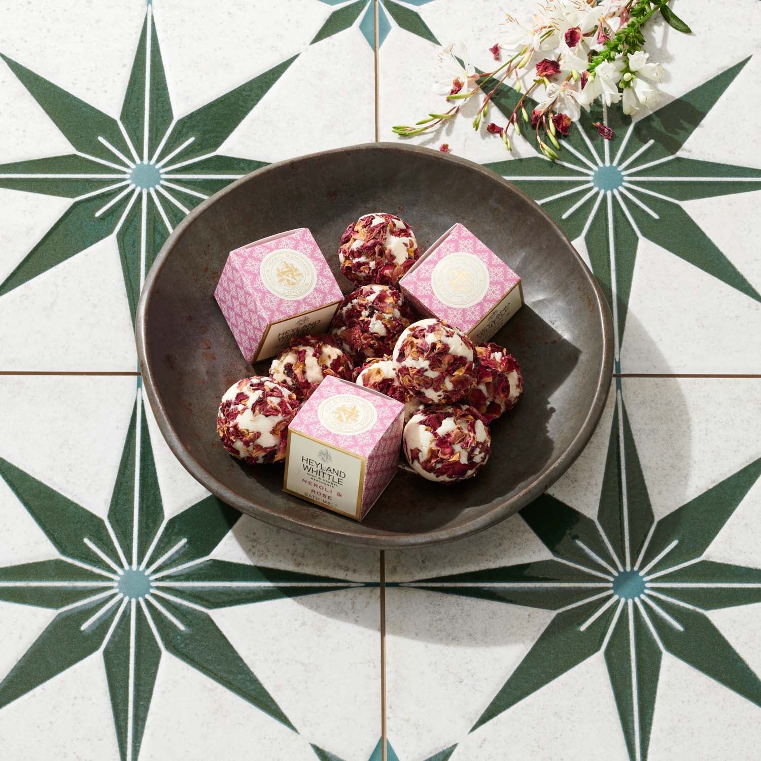 A dark bowl filled with pink and white packaged bath melts, arranged on a patterned tile surface with decorative flowers in the corner.