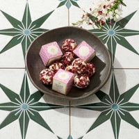 A dark bowl filled with pink and white packaged bath melts, arranged on a patterned tile surface with decorative flowers in the corner.