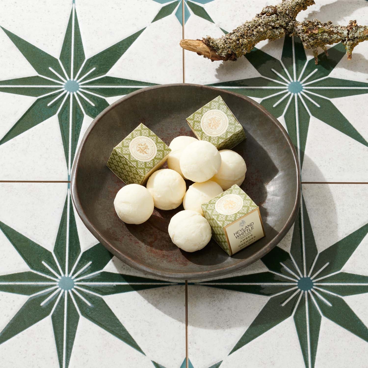 A dark ceramic bowl filled with round white bath melts and two small patterned boxes, arranged on a decorative tile surface with a branch in the corner.