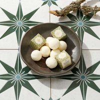 A dark ceramic bowl filled with round white bath melts and two small patterned boxes, arranged on a decorative tile surface with a branch in the corner.