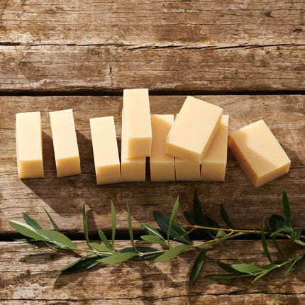 Several pale yellow rectangular bars of soap arranged on a rustic wooden surface, with green olive branches placed along the bottom of the frame.