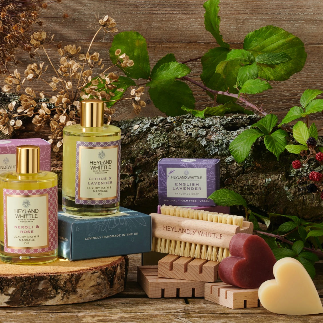 A styled product display of Heyland Whittle bath and body items on a rustic wooden surface, including two small bottles with gold caps, two purple boxes, two wooden hand brushes, two heart-shaped soap bars, dried flowers, green leaves, and a log background.
