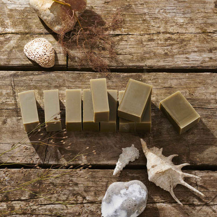 Several rectangular bars of handmade soap in muted olive green are arranged on rustic wooden planks, styled with seashells, dried grass, and stones.