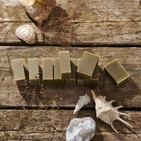 Several rectangular bars of handmade soap in muted olive green are arranged on rustic wooden planks, styled with seashells, dried grass, and stones.