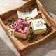 A luxury gift box set from Heyland Whittle, featuring a gold-toned outer box with a cream lid and a pink-lined inner box containing assorted chocolates, displayed on a rustic wooden tray with rosebuds.