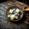 A dark ceramic bowl filled with bath melts and two light green square boxes, set on a rustic wooden table with a small lit candle in the background.