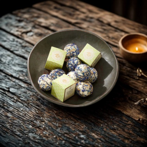 A dark ceramic bowl filled with bath melts and two light green square boxes, set on a rustic wooden table with a small lit candle in the background.