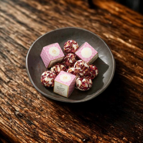 A dark gray ceramic bowl filled with pink and white patterned square boxes and several round pink-and-white marbled bath melts, set on a rustic dark wooden tabletop.