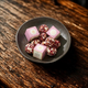 A dark gray ceramic bowl filled with pink and white patterned square boxes and several round pink-and-white marbled bath melts, set on a rustic dark wooden tabletop.