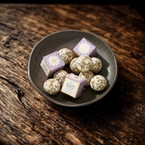 Three small lavender and white boxes with bath melts arranged in a dark ceramic bowl on a rustic wooden surface.