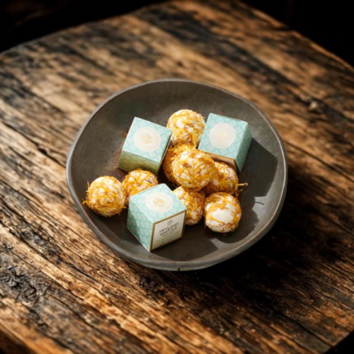 Four small square boxes with a light blue-green patterned design and white circular labels, alongside several bath melts, arranged in a dark gray ceramic bowl on a rustic wooden surface.