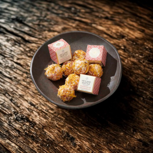 A dark ceramic plate holding several bath melts, with two small pink-and-white branded boxes placed on top, set on a rustic dark wooden surface.