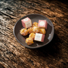 A dark ceramic plate holding several bath melts, with two small pink-and-white branded boxes placed on top, set on a rustic dark wooden surface.